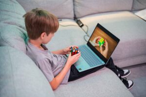 Young boy sitting on a couch solving a Rubik’s cube while watching an instructional video on a laptop.