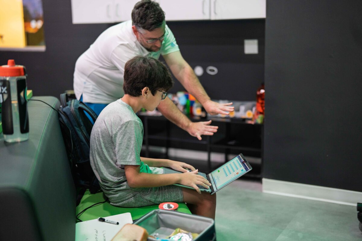 Boy using a laptop while an adult coach or mentor gestures toward the screen, providing guidance in a learning or coding session.