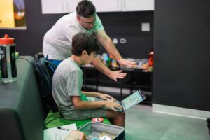 Boy using a laptop while an adult coach or mentor gestures toward the screen, providing guidance in a learning or coding session.