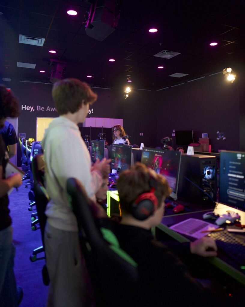 A group of students participating in a gaming session at NextGen Academy. The room is dimly lit with purple overhead lighting, and multiple computer monitors display a video game. A student in the foreground is seated with headphones on, focusing on the screen, while others stand and observe. The background features a wall with the text “Hey, Be Awesome Today!"