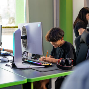 Smiling student writes in a notebook at a computer desk with a gaming PC, mechanical keyboard, and headphones nearby.